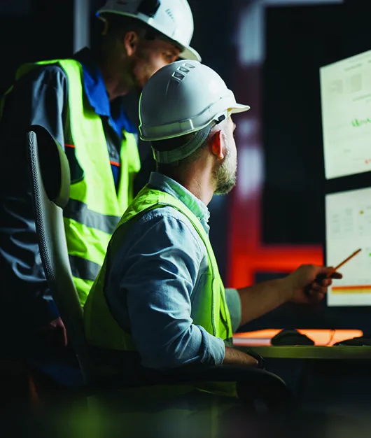 AI in industrials: Two men in high viz jackets and hard hats looking at a bank of screens